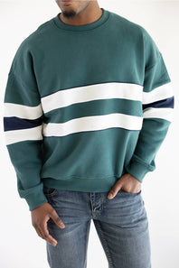 A man wearing a dark teal crewneck sweatshirt with horizontal white and navy blue stripes across the chest and sleeves, paired with blue jeans.