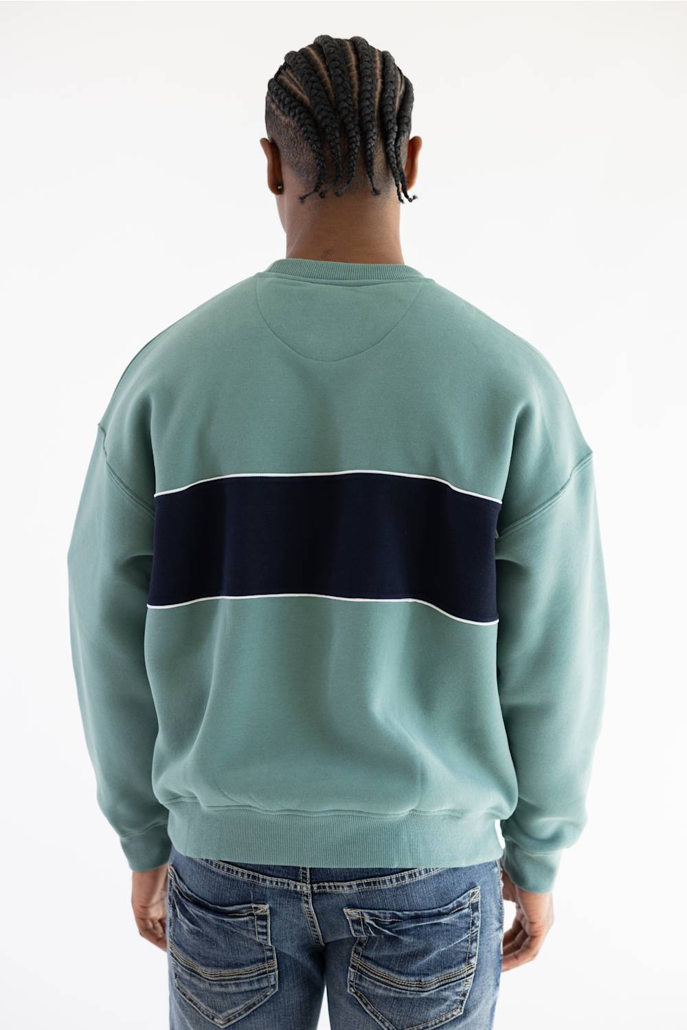 A man viewed from behind wearing a sage green crewneck sweatshirt with a wide navy blue horizontal stripe across the back, paired with blue jeans, against a plain white background.