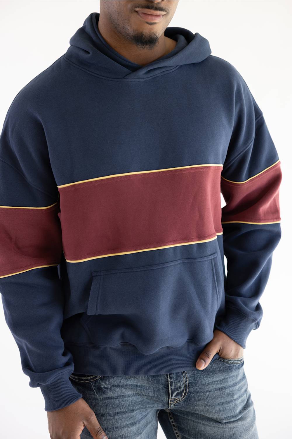 A man wearing a navy blue hoodie with a wide burgundy horizontal stripe across the chest and sleeves, outlined with thin gold piping, paired with blue jeans against a plain white background.