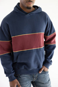 A man wearing a navy blue hoodie with a wide burgundy horizontal stripe across the chest and sleeves, outlined with thin gold piping, paired with blue jeans against a plain white background.