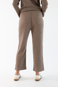 A person shown from the back wearing a ribbed knit sweater and matching cropped ribbed lounge pants in a sand-brown color, paired with white sneakers, against a plain white studio background.