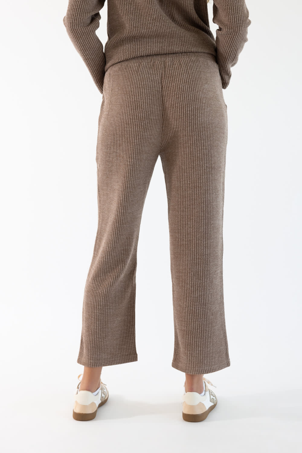 A person shown from the back wearing a ribbed knit sweater and matching cropped ribbed lounge pants in a sand-brown color, paired with white sneakers, against a plain white studio background.