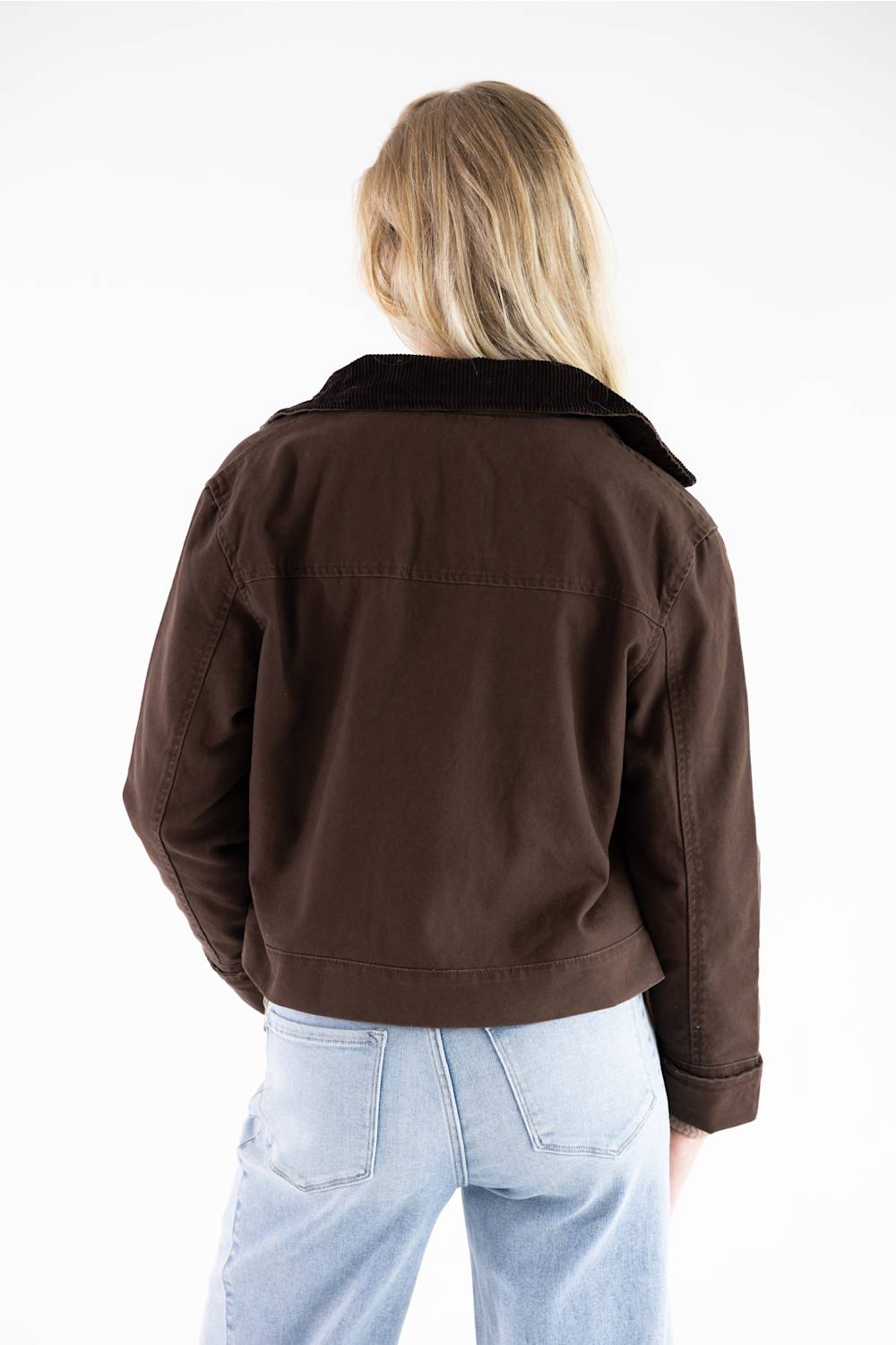 A woman viewed from behind wearing a dark brown cropped jacket with a black collar and light blue jeans, set against a plain white studio background.