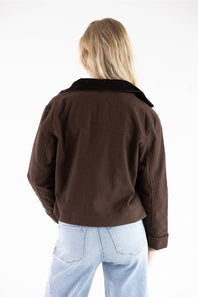 A woman viewed from behind wearing a dark brown cropped jacket with a black collar and light blue jeans, set against a plain white studio background.
