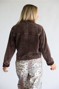 A woman viewed from the back wearing a dark brown corduroy jacket and patterned patchwork pants against a light gray studio background.