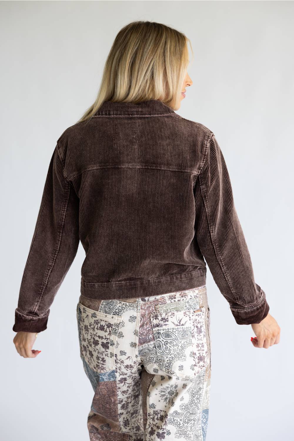 A woman viewed from the back wearing a dark brown corduroy jacket and patterned patchwork pants against a light gray studio background.