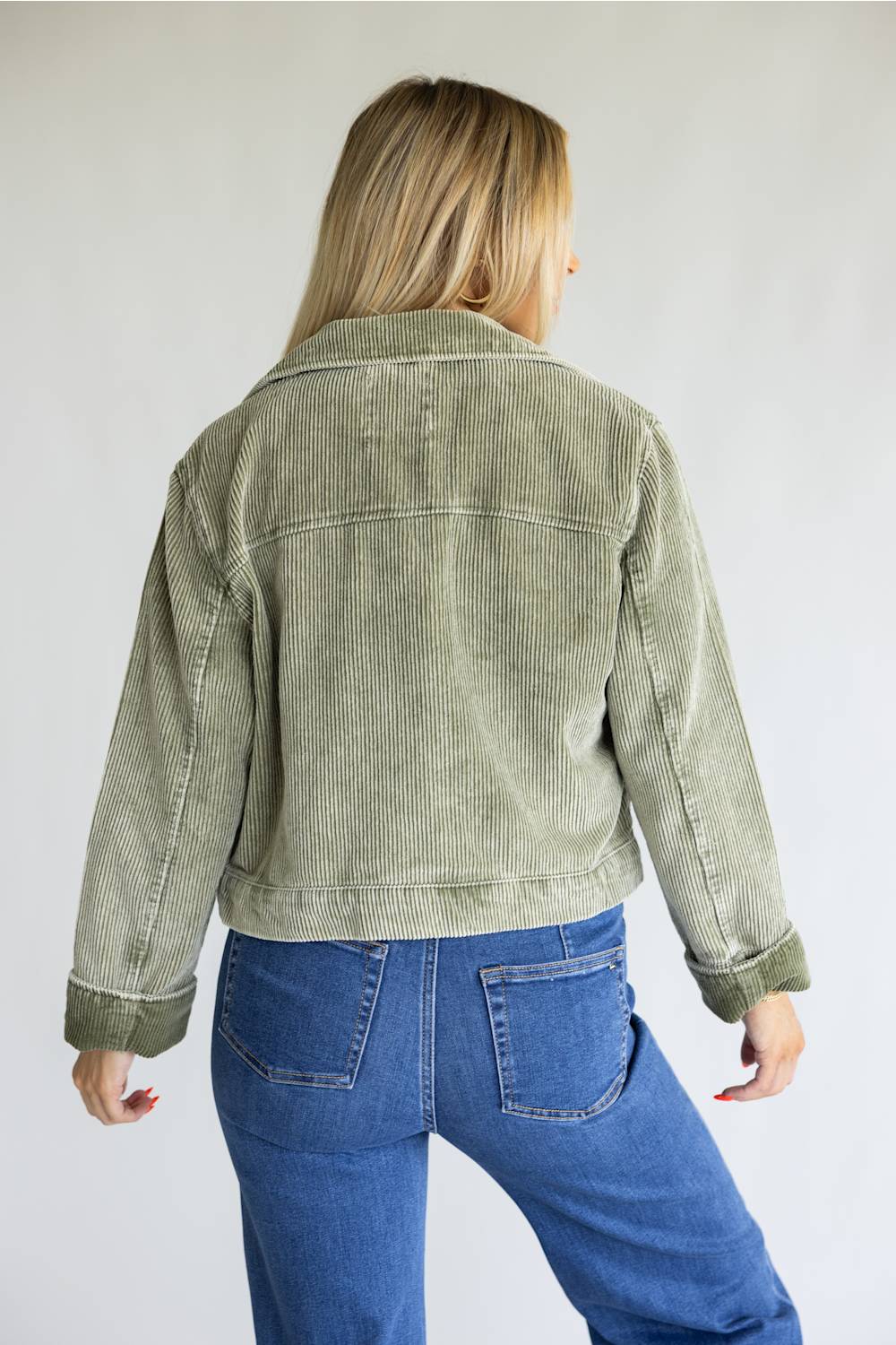 A woman viewed from behind wearing a cropped sage green corduroy jacket and blue jeans, standing against a plain light gray studio background.