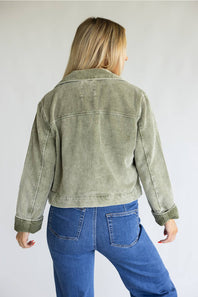 A woman viewed from behind wearing a cropped sage green corduroy jacket and blue jeans, standing against a plain light gray studio background.