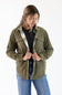 A woman wearing an olive green corduroy button-up shirt jacket over a black top and blue jeans, standing against a plain white background.