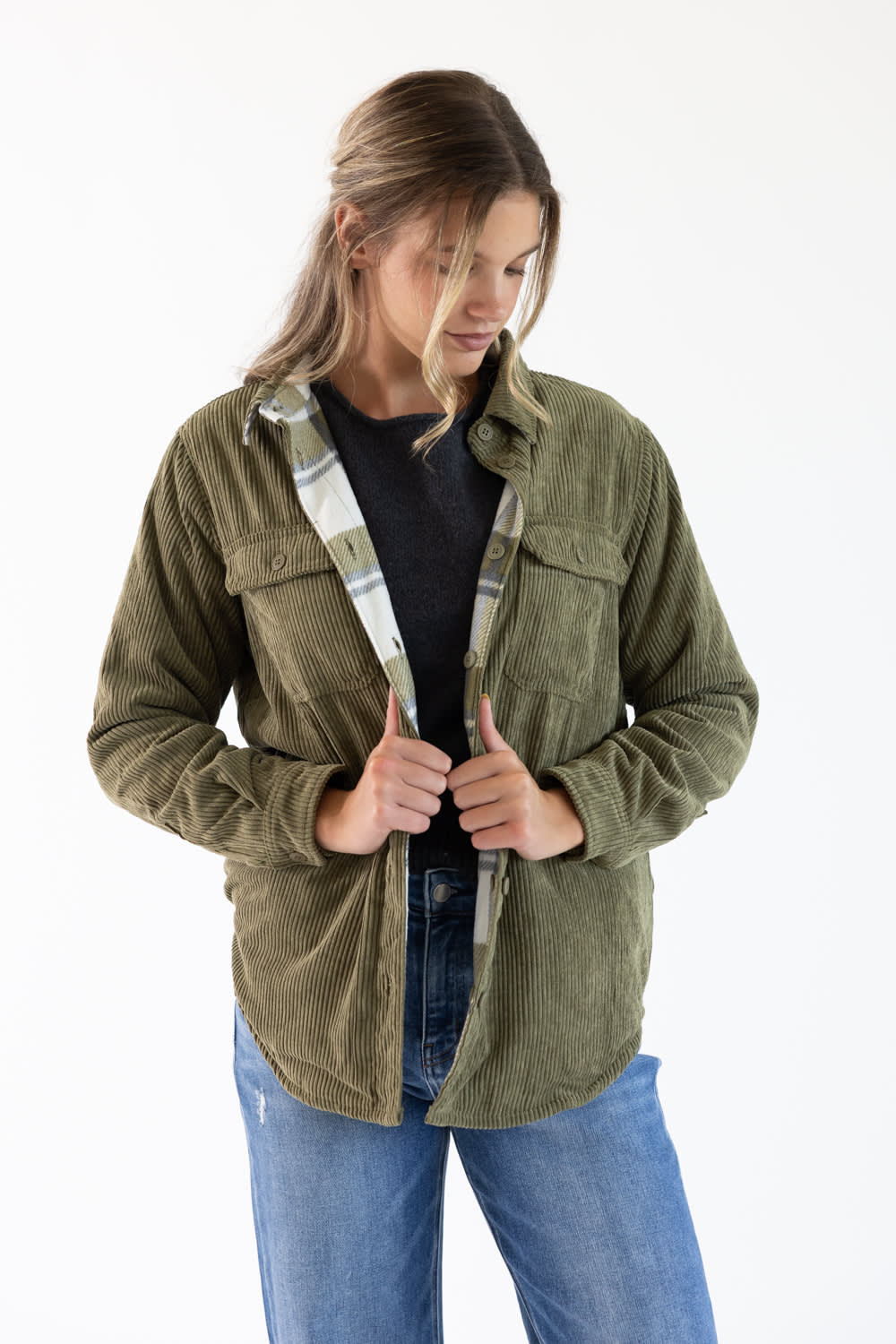 A woman wearing an olive green corduroy button-up shirt jacket over a black top and blue jeans, standing against a plain white background.