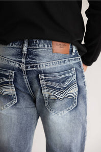 Close-up back view of a person wearing light-to-medium blue denim jeans with decorative contrast stitching and a tan leather patch reading DAKOTA JEANS, paired with a black long-sleeve top against a plain light background.