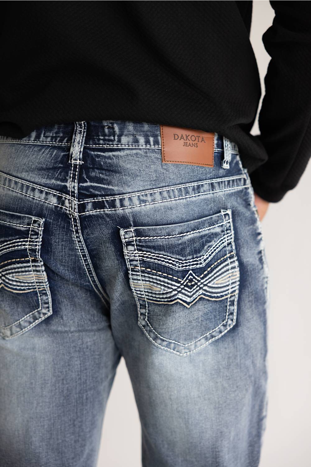 Close-up back view of a person wearing light-to-medium blue denim jeans with decorative contrast stitching and a tan leather patch reading DAKOTA JEANS, paired with a black long-sleeve top against a plain light background.