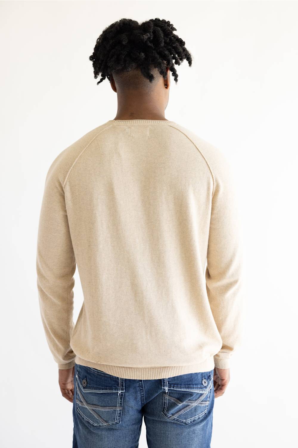 A man viewed from behind wearing a light beige crewneck sweater and blue jeans against a plain white background.