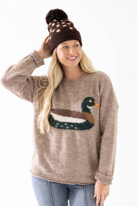 A woman wearing a brown knit beanie with a pom-pom and a taupe sweater featuring a large embroidered duck design on the front, paired with blue jeans, against a plain white background.
