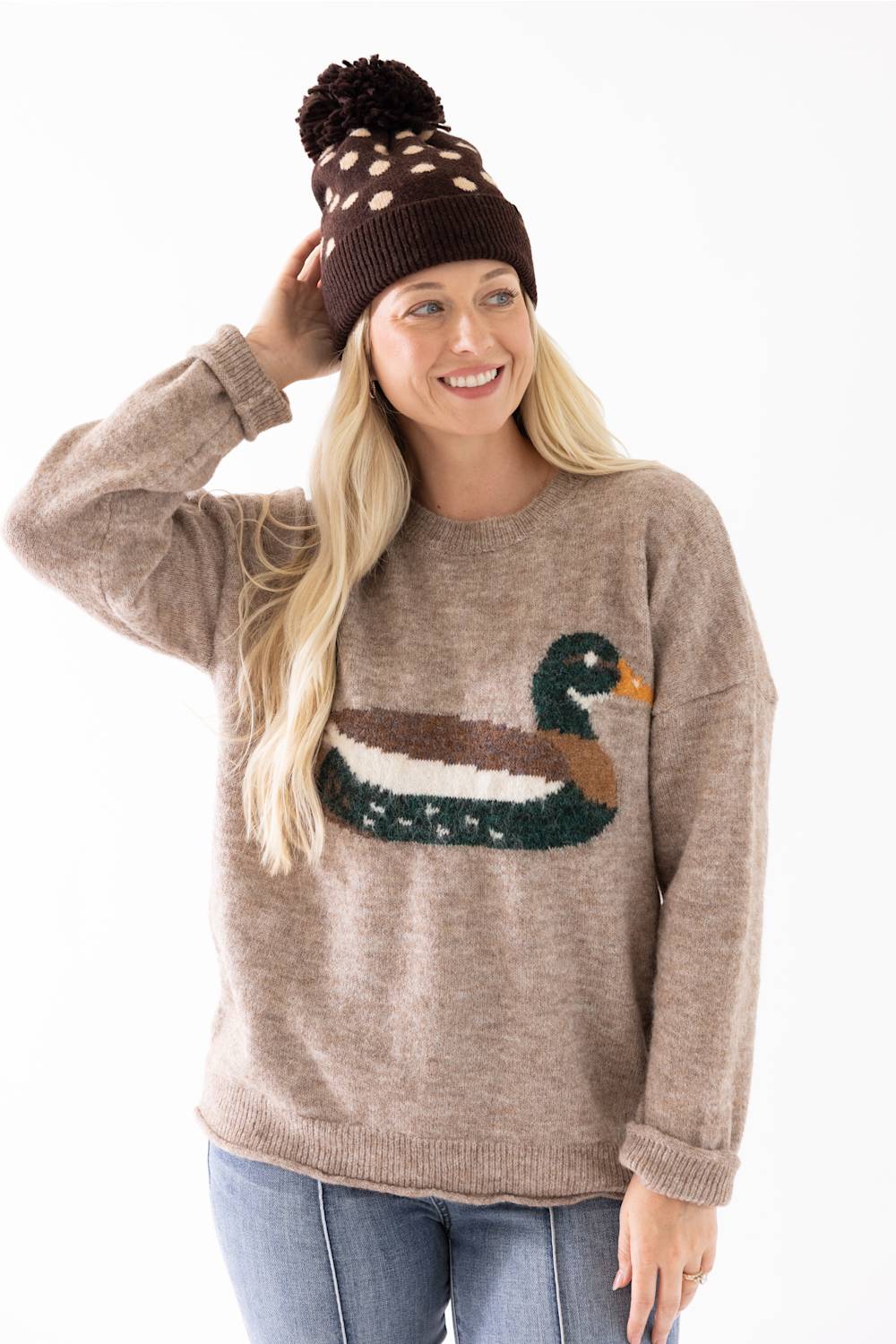 A woman wearing a brown knit beanie with a pom-pom and a taupe sweater featuring a large embroidered duck design on the front, paired with blue jeans, against a plain white background.