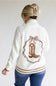 A woman shown from the back wearing a cream knit cardigan with a brown embroidered cowboy boot and bow design on the back, paired with blue jeans against a plain light gray background.
