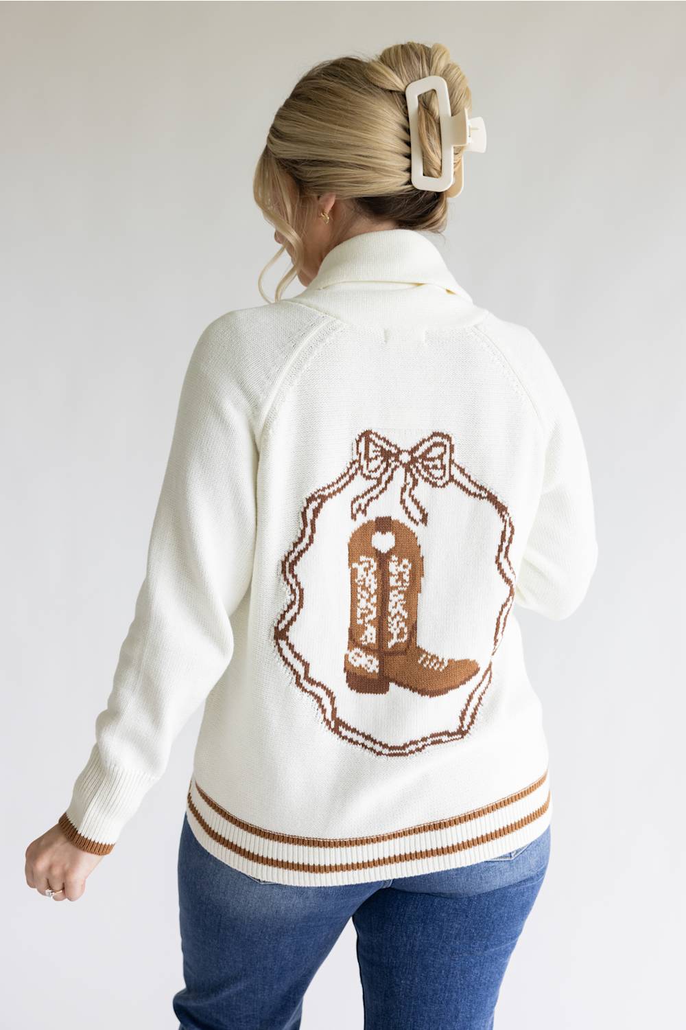 A woman shown from the back wearing a cream knit cardigan with a brown embroidered cowboy boot and bow design on the back, paired with blue jeans against a plain light gray background.