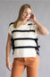A woman modeling a cream knit short-sleeve sweater with bold black horizontal stripes and side tie details, paired with rust-orange pants, against a solid gray studio background.