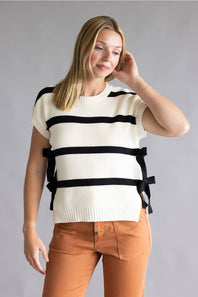 A woman modeling a cream knit short-sleeve sweater with bold black horizontal stripes and side tie details, paired with rust-orange pants, against a solid gray studio background.