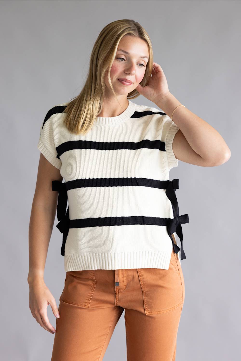 A woman modeling a cream knit short-sleeve sweater with bold black horizontal stripes and side tie details, paired with rust-orange pants, against a solid gray studio background.