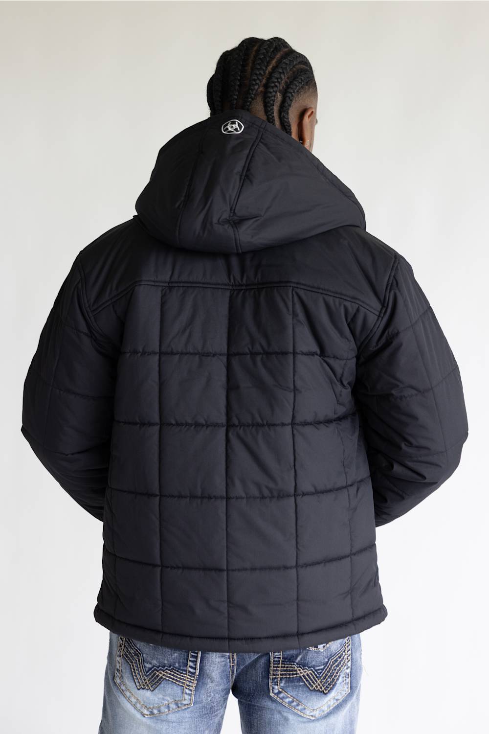A man viewed from behind wearing a black quilted puffer jacket with a hood, paired with blue jeans, against a plain light gray background.