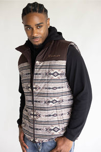 A man wearing a brown and patterned zip-up vest over a black hoodie, standing against a plain light background.