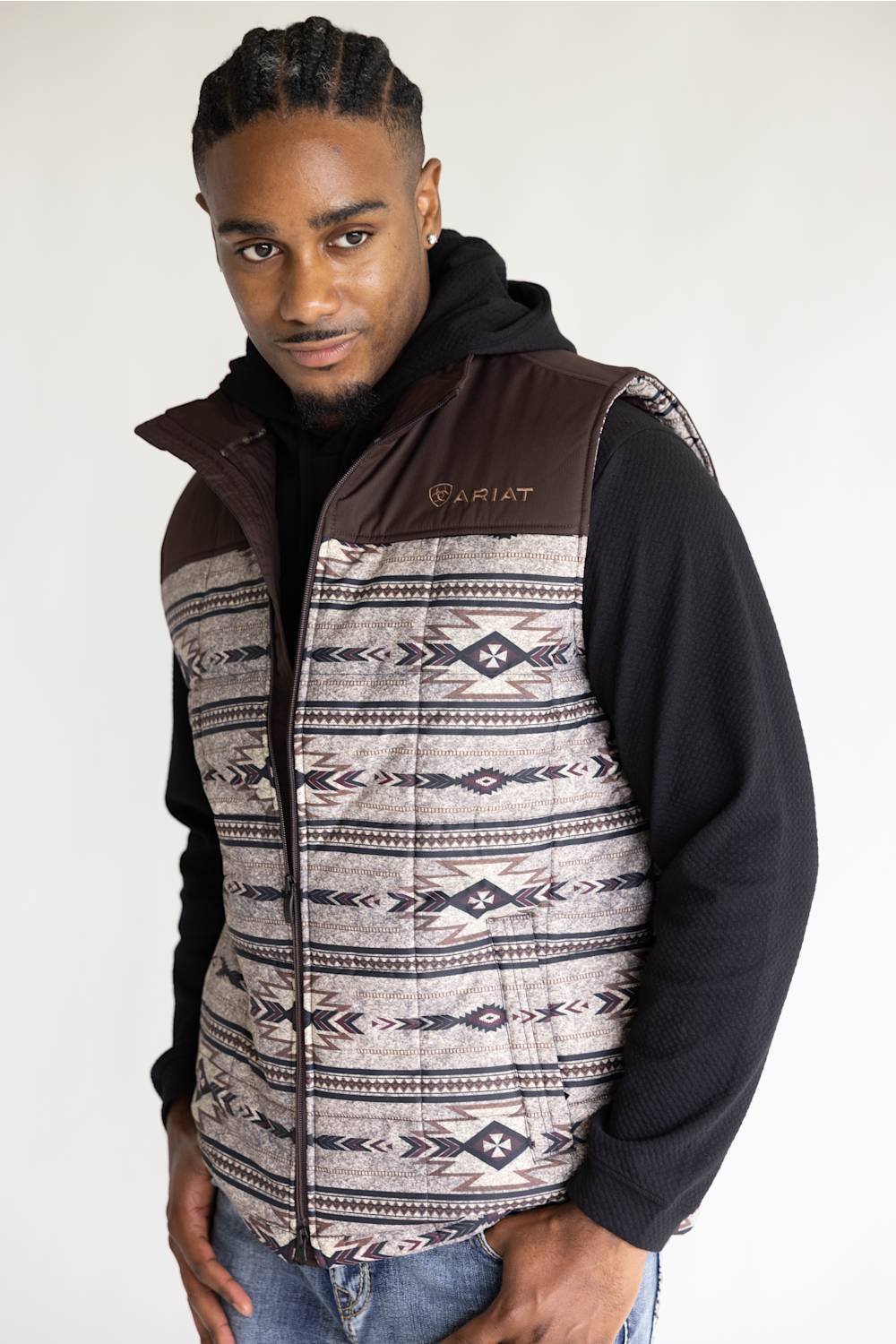 A man wearing a brown and patterned zip-up vest over a black hoodie, standing against a plain light background.