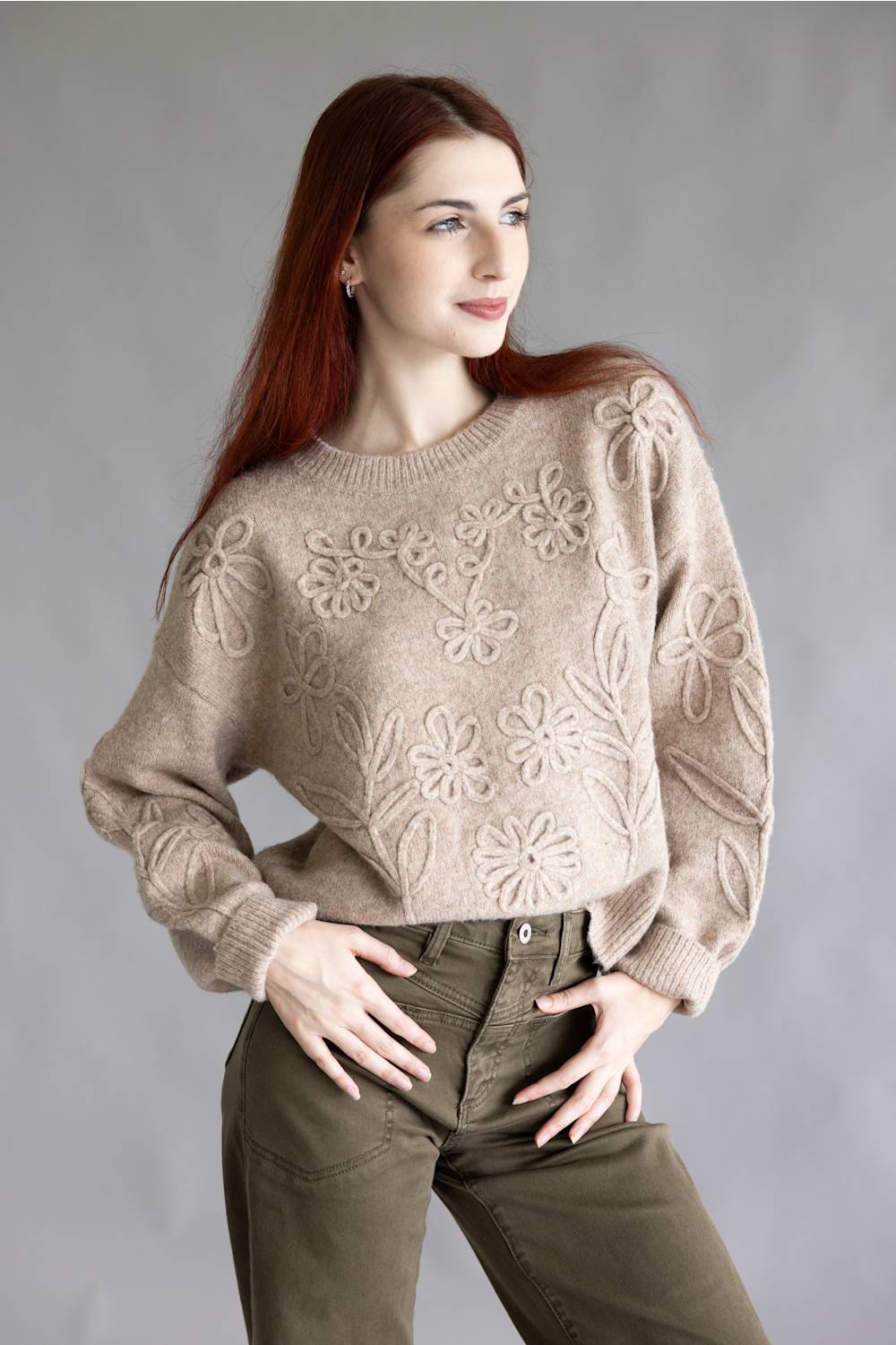 A woman with long red hair wearing a beige knit sweater with raised floral patterns and olive green pants, posing against a neutral gray studio background.
