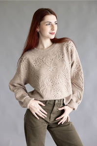 A woman with long red hair wearing a beige knit sweater with raised floral patterns and olive green pants, posing against a neutral gray studio background.