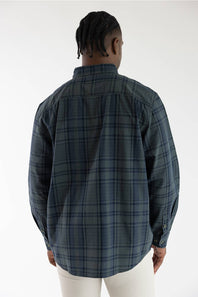 Back perspective of a male model wearing the long-sleeve Carhartt Fort Loose Plaid Shirt. The shirt has a navy and green plaid pattern. He is wearing it with light-colored pants.