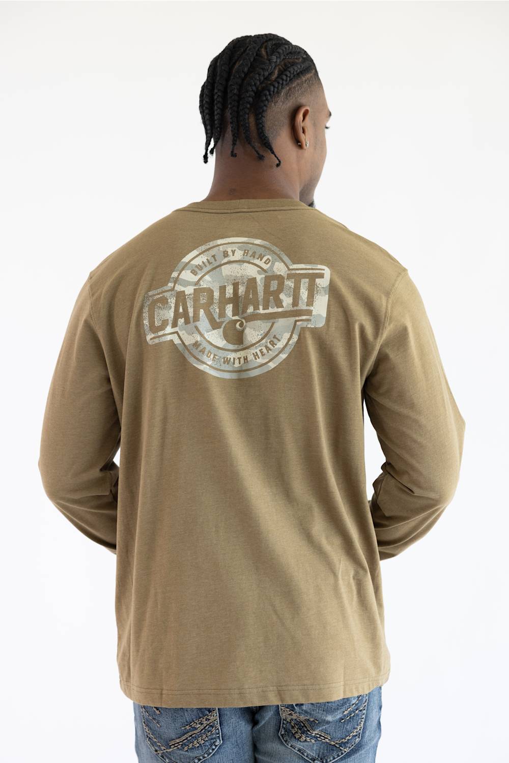 A man viewed from the back wearing a long sleeve light brown t-shirt with a graphic that says "CARHARTT BUILD BY HAND MADE WITH HEART" with a Carhartt logo C.