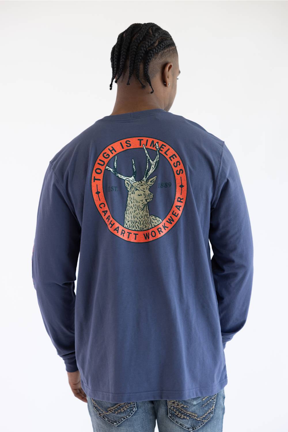 A man viewed from behind wearing a navy blue long-sleeve t-shirt with a large circular back graphic featuring a deer head and the text 'TOUGH IS TIMELESS' and 'CARHARTT WORKWEAR'.