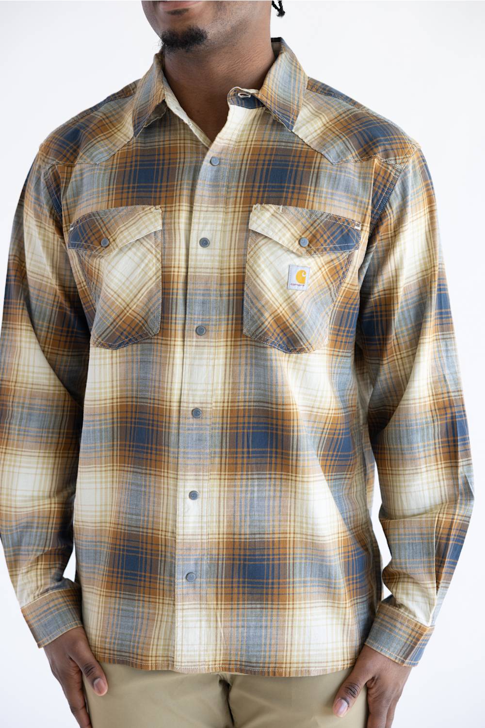 A close up of a man wearing a Carhartt long-sleeve plaid button-up shirt in beige, brown, and blue tones, paired with khaki pants, against a plain white background.