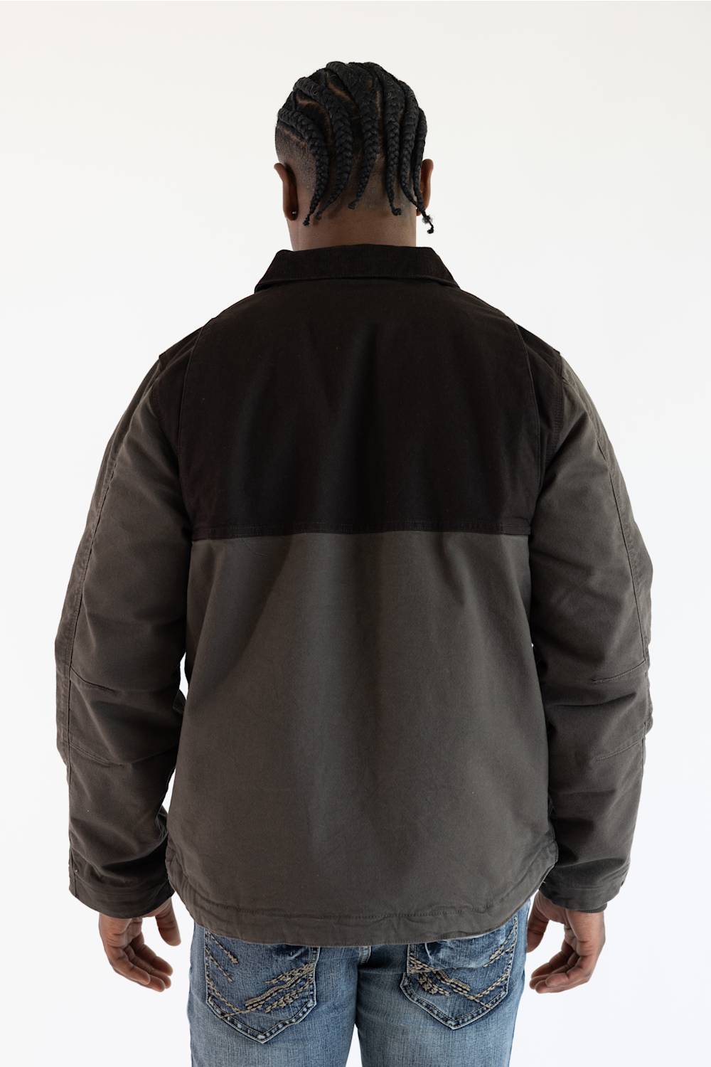A person with braided hair stands facing away from the camera, wearing a two-toned jacket and jeans against a plain white background.