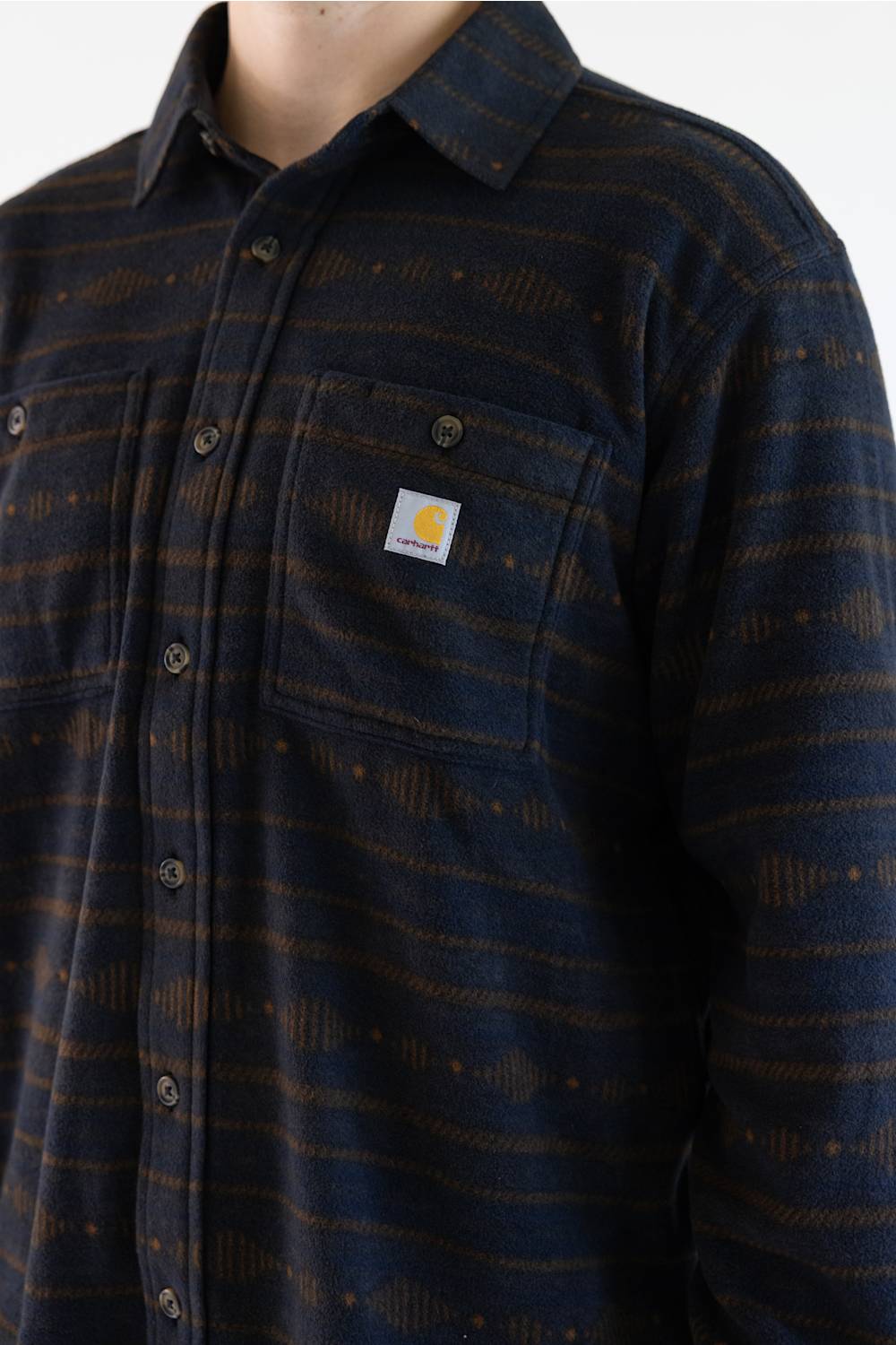 A man wearing a long-sleeve Carhartt plaid button-up shirt in navy and brown with thin orange stripes, paired with blue jeans, against a plain white background.