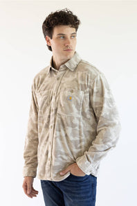 A man wearing a beige and off-white camouflage Carhartt long-sleeve button-up shirt with blue jeans, standing against a plain white studio background.