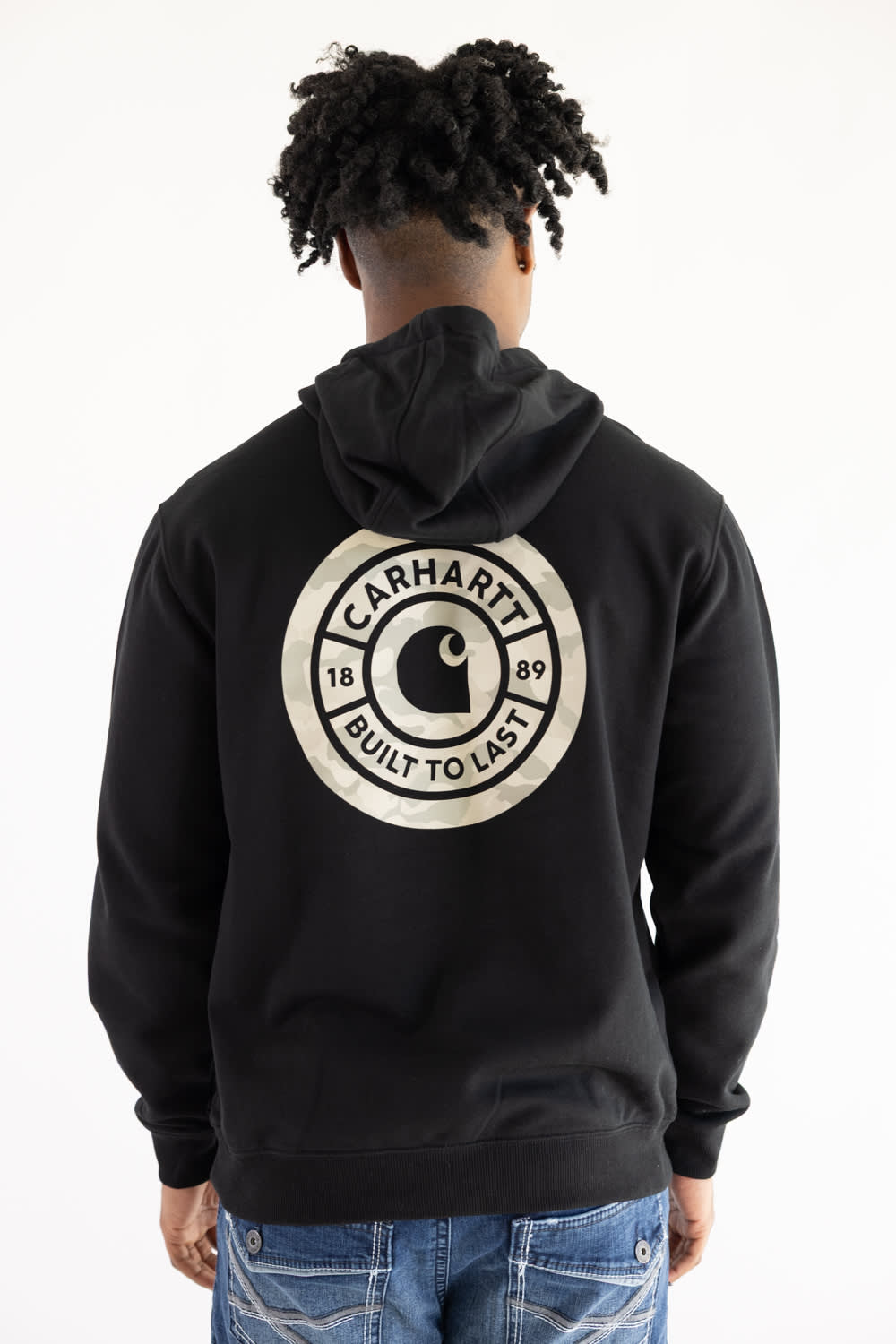Back view of a man wearing a black Carhartt Sawyer fleece hoodie with a large circular emblem graphic featuring camo print, the Carhartt "C" logo, and "1889 Built To Last" lettering.