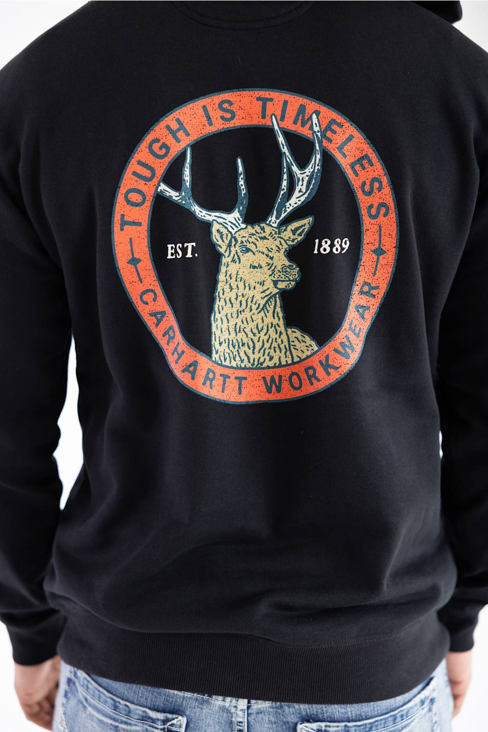 Back view of a man wearing a black Carhartt hooded sweatshirt featuring a large circular graphic on the back. The graphic displays a distressed orange border with the text "TOUGH IS TIMELESS" and "CARHARTT WORKWEAR," enclosing an illustration of a buck deer, the text "EST. 1889," and decorative stars.