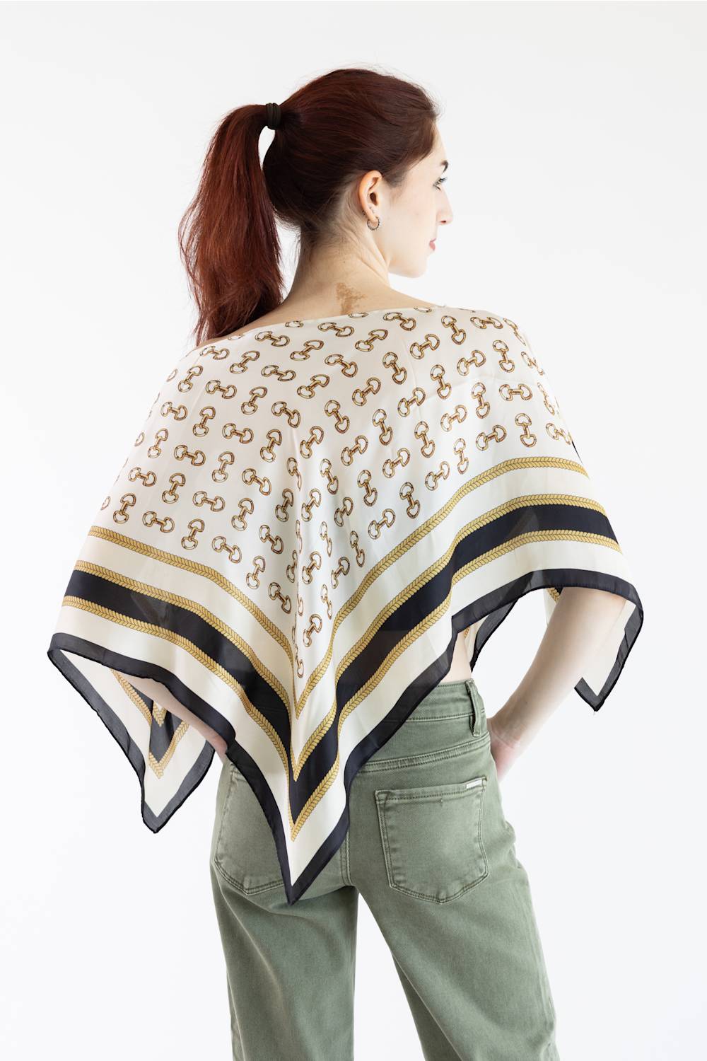 A woman shown from the back wearing a white scarf with a repeating brown monogram pattern and black and gold striped borders, paired with sage green pants against a white studio background.