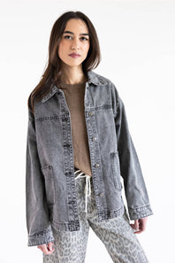 A woman modeling a washed gray denim jacket over a brown top and leopard-print pants against a plain white background.