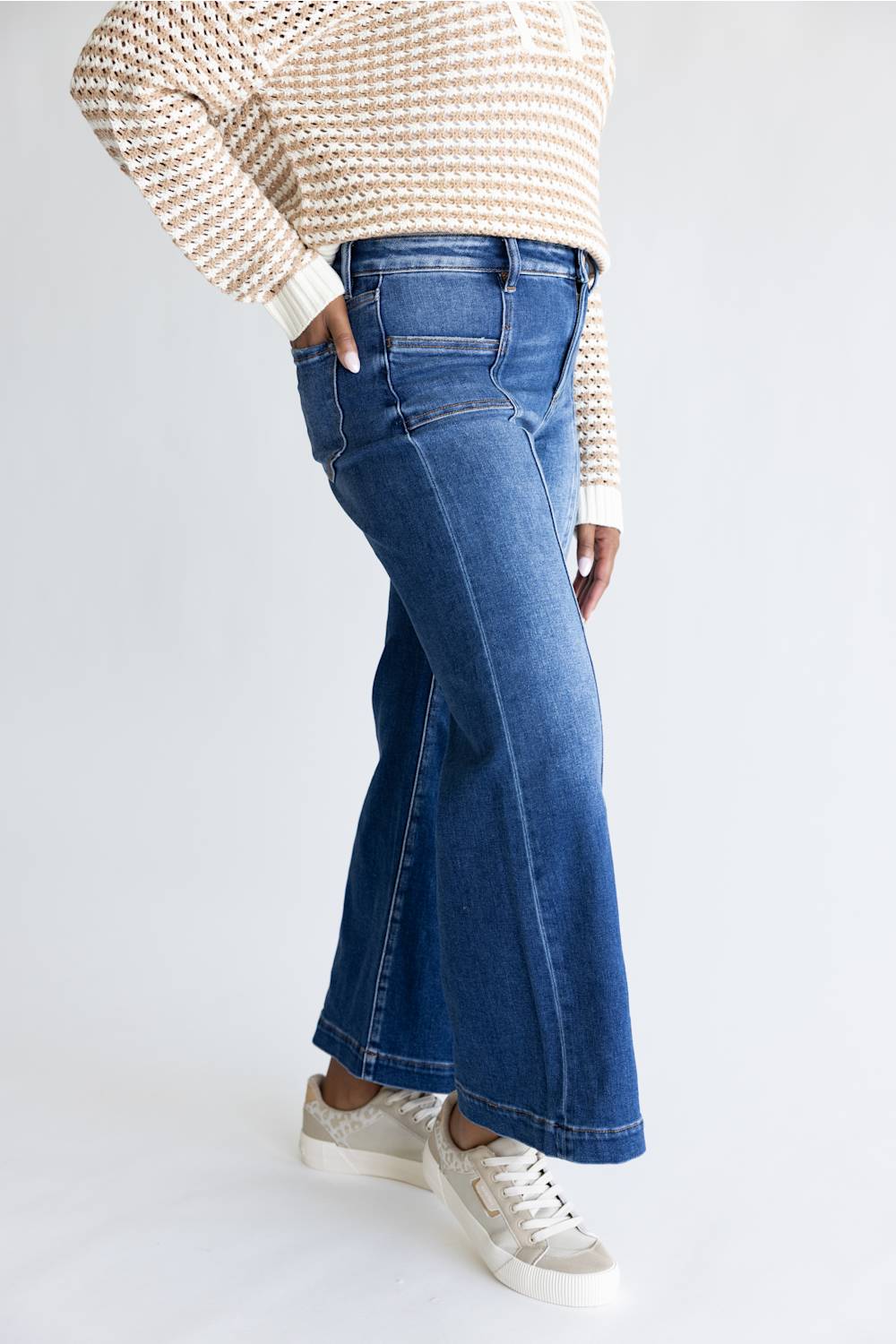 A person wearing blue wide-leg cropped jeans, a beige and white patterned knit sweater, and white sneakers, shown from the chest down against a light gray studio background.
