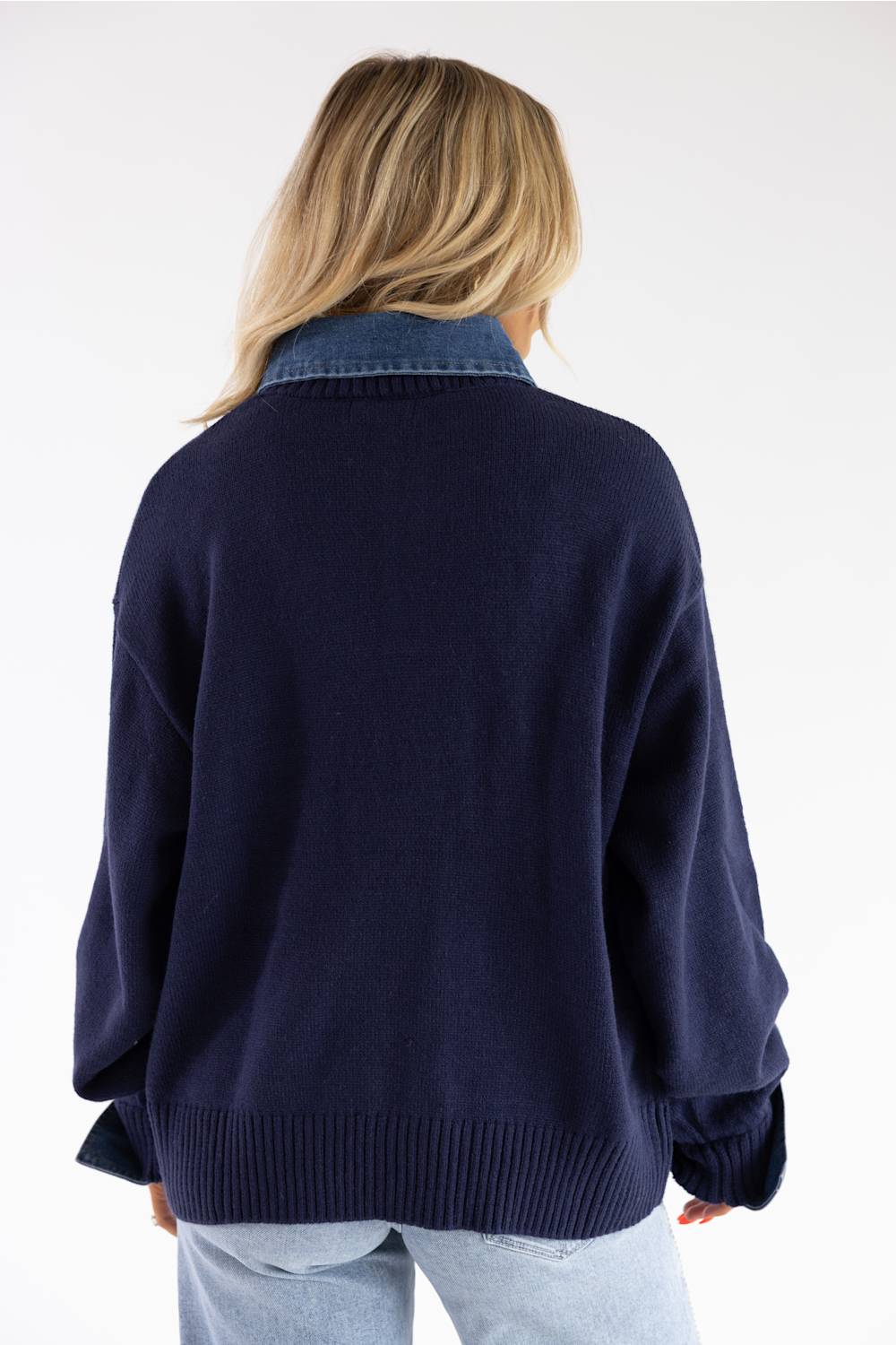 A woman viewed from behind wearing a navy blue knit cardigan with a contrasting blue denim collar, paired with light blue jeans, against a plain white background.