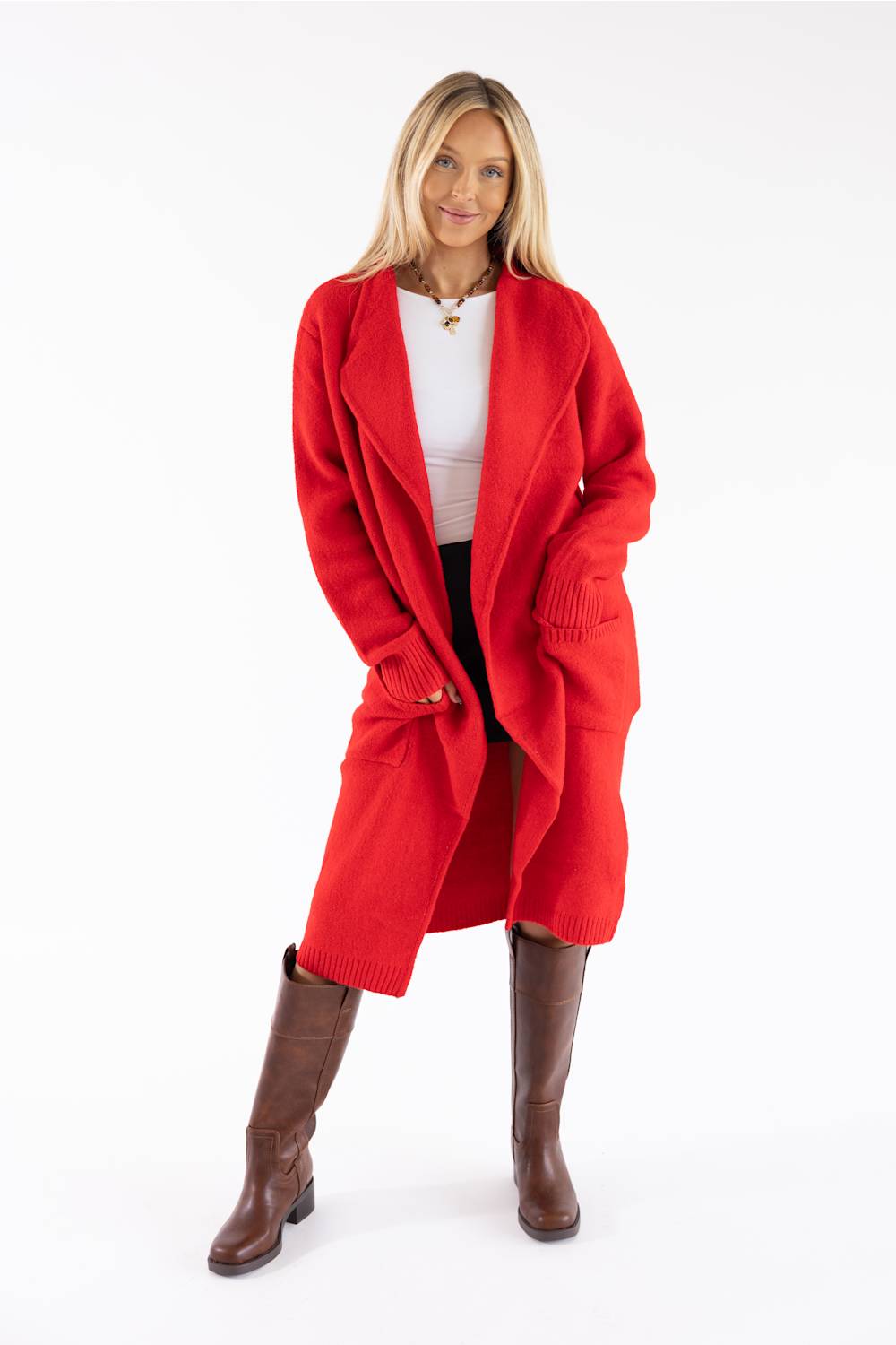 A woman modeling a bright red open-front knit cardigan over a white top and black pants, paired with brown knee-high boots, against a plain white studio background.