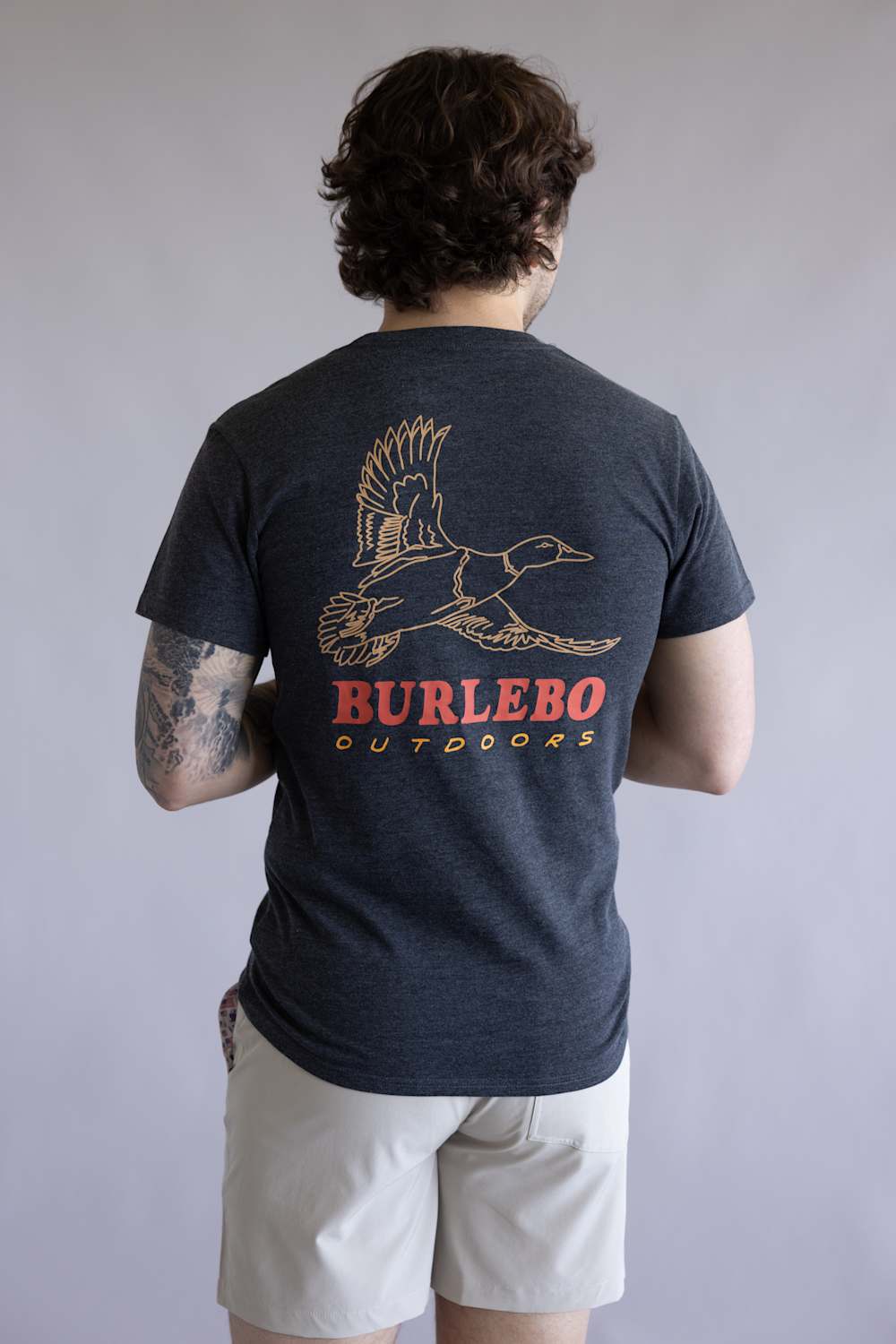 Back view of a person wearing a navy t-shirt with a bird graphic and 'Burlebo Outdoors' text on the back, standing against a plain background. The shirt is Burlebo Duck Wild Tee for Men in Heather Black
