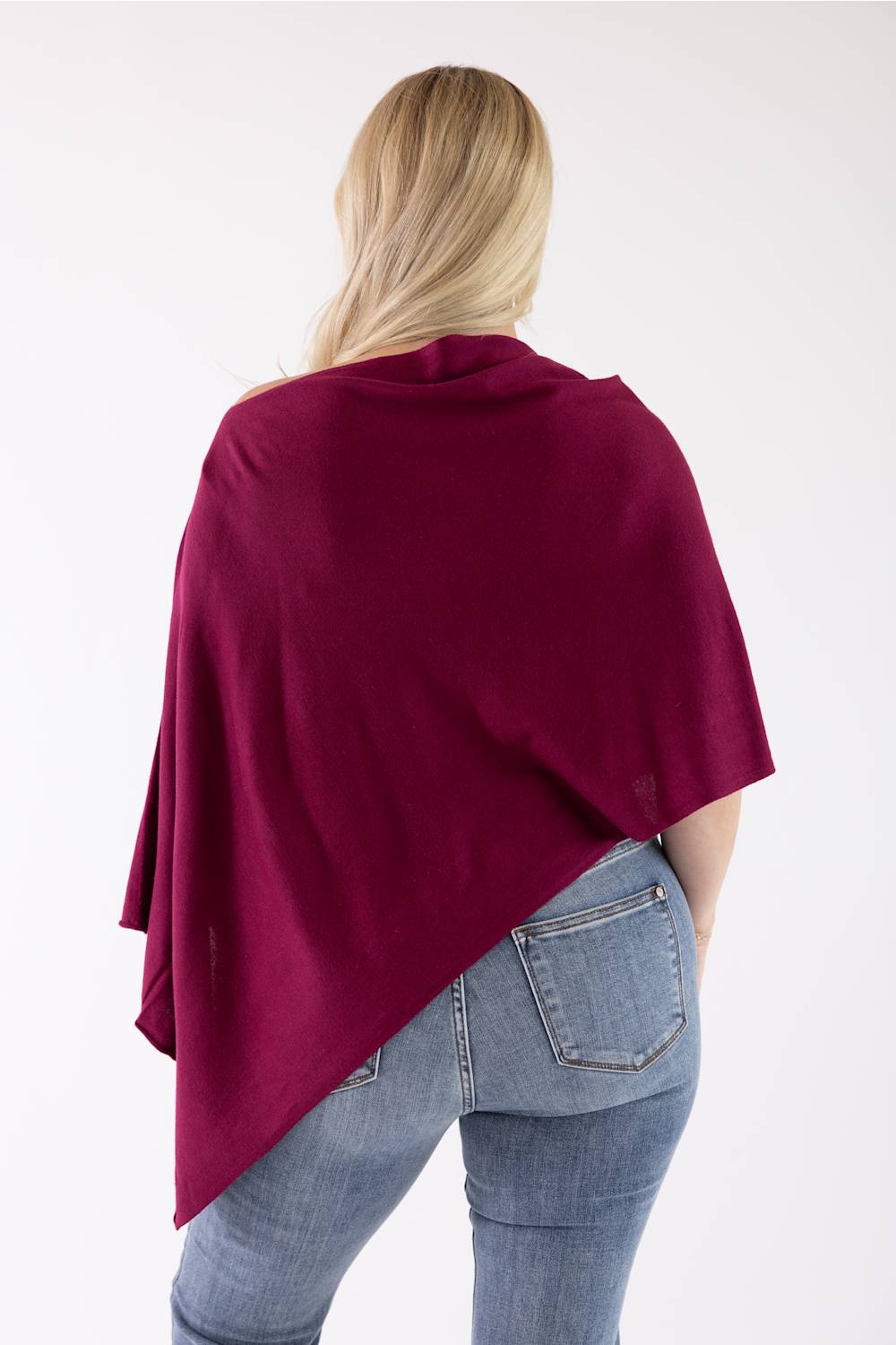 A woman viewed from behind wearing a deep burgundy draped wrap or poncho over light blue jeans, set against a plain white studio background.