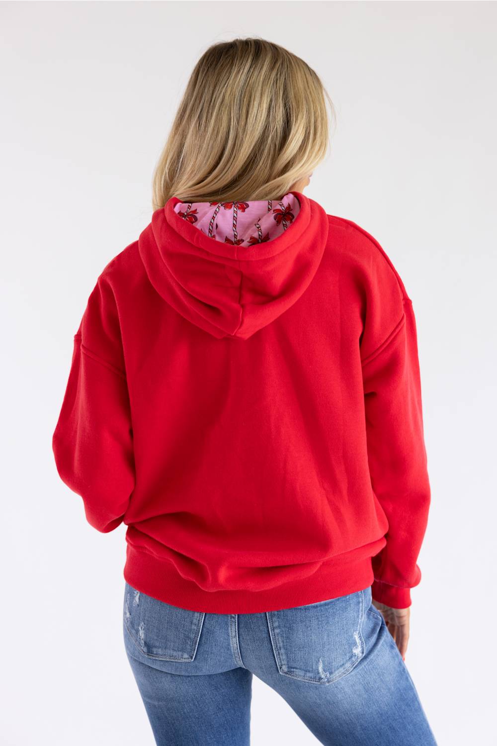 A woman viewed from behind wearing a bright red pullover hoodie with a pink floral patterned lining inside the hood, paired with blue jeans against a plain white background.