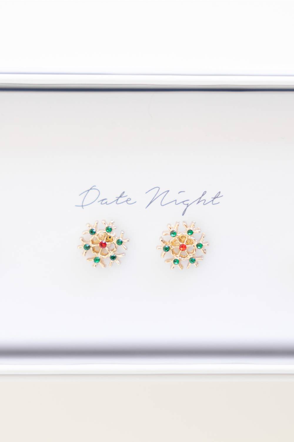 A pair of gold-toned floral stud earrings with green and red stones, displayed on a white card labeled 'Date Night' inside a white box.