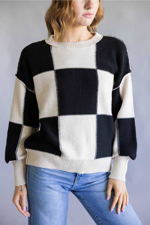 A woman wearing a black and cream checkered knit sweater with blue jeans, shown from the neck down against a neutral gray background.