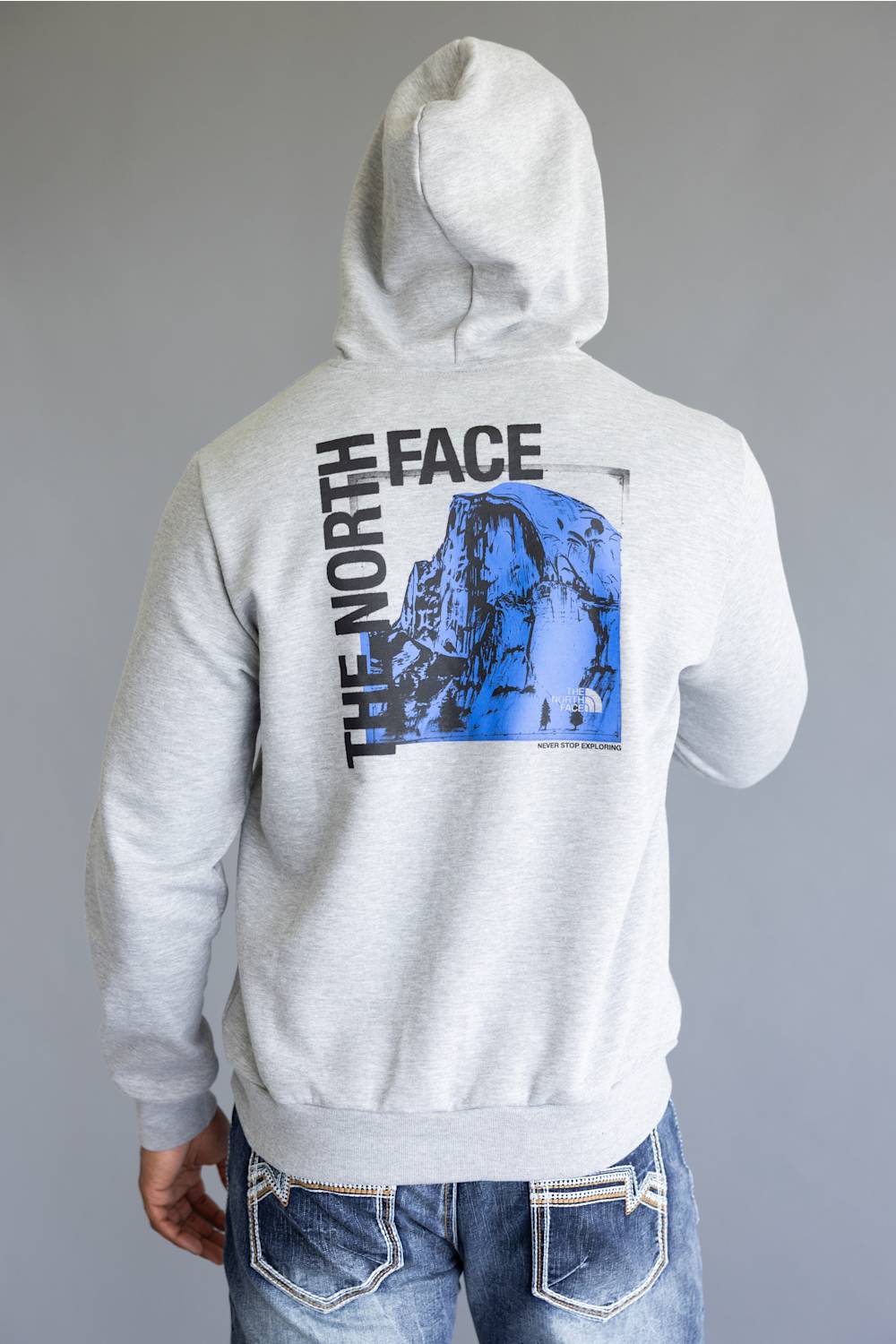 A person shown from the back wearing a light heather gray hoodie with a large black and blue graphic print on the back, paired with blue jeans, against a plain gray studio background.