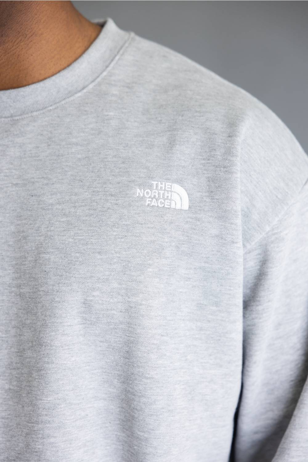 Close-up of a person wearing a light heather gray crewneck sweatshirt with a small white embroidered The North Face logo on the left chest, set against a neutral gray background.
